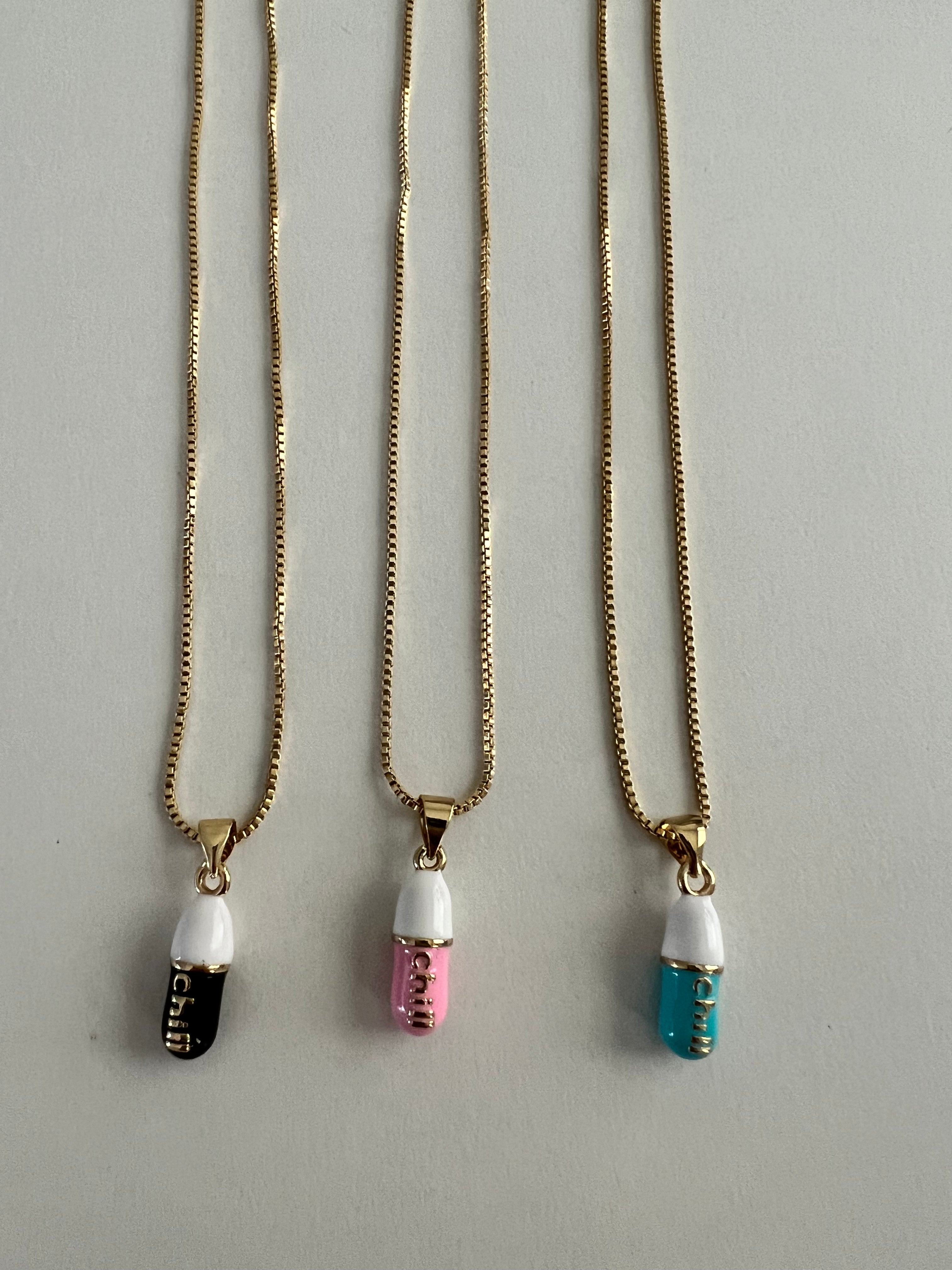 three separate gold chain pendants holding a pink, tiffany blue and black colored pill charm with the word chill written in gold letters on the pill charm