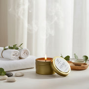 Gold candle tin with a white candle inside on a textured white surface surrounded by towels and rocks in a spa-like setting