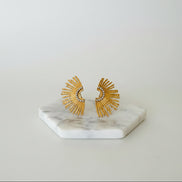 A pair of gold-colored sunburst earrings displayed on a white marble hexagonal stand.