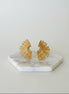 A pair of gold-colored sunburst earrings displayed on a white marble hexagonal stand.