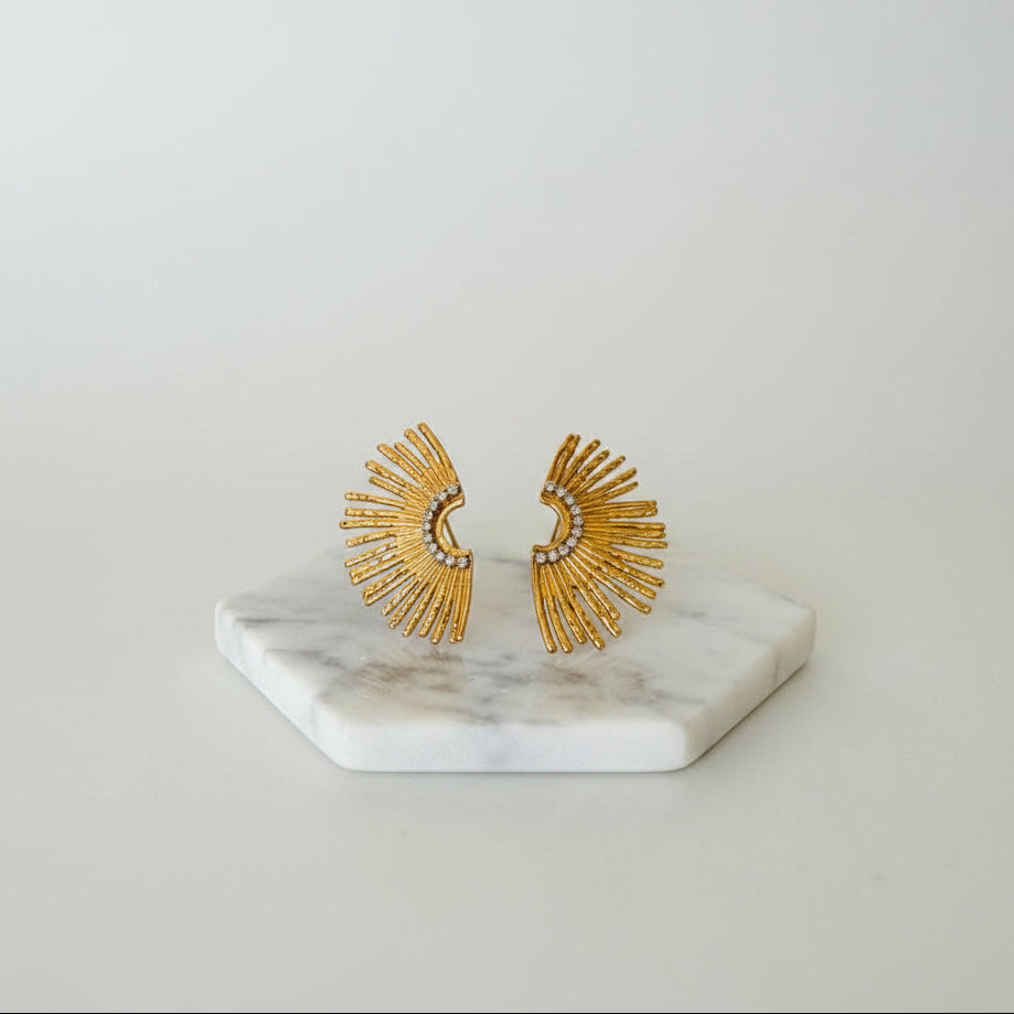A pair of gold-colored sunburst earrings displayed on a white marble hexagonal stand.